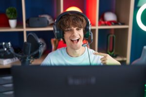 Young man wearing headphones, smiling at a computer, pointing left — representing webcam modeling at home