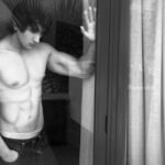 Black and white portrait of a shirtless man standing beside a window curtain