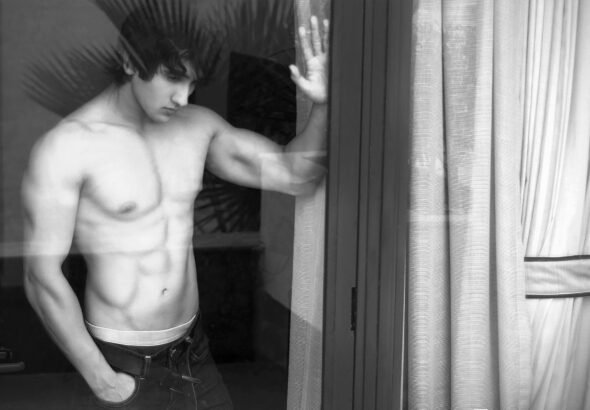 Black and white portrait of a shirtless man standing beside a window curtain