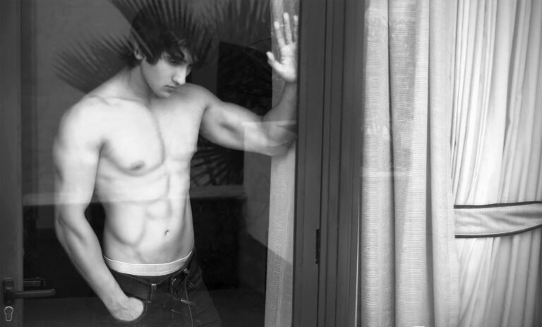 Black and white portrait of a shirtless man standing beside a window curtain